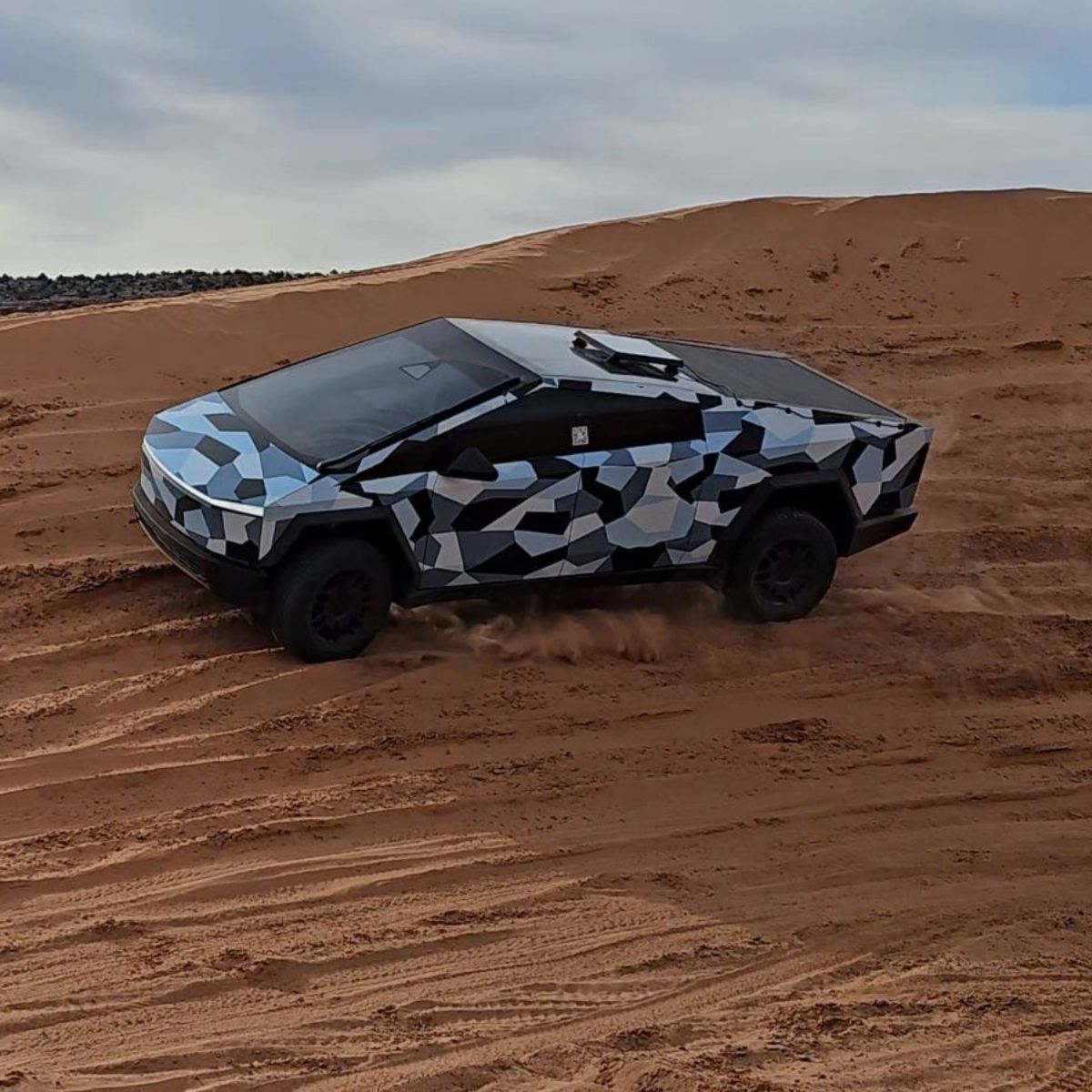Cybertruck wrapped in a grey and black camouflage livery driving on sand dunes