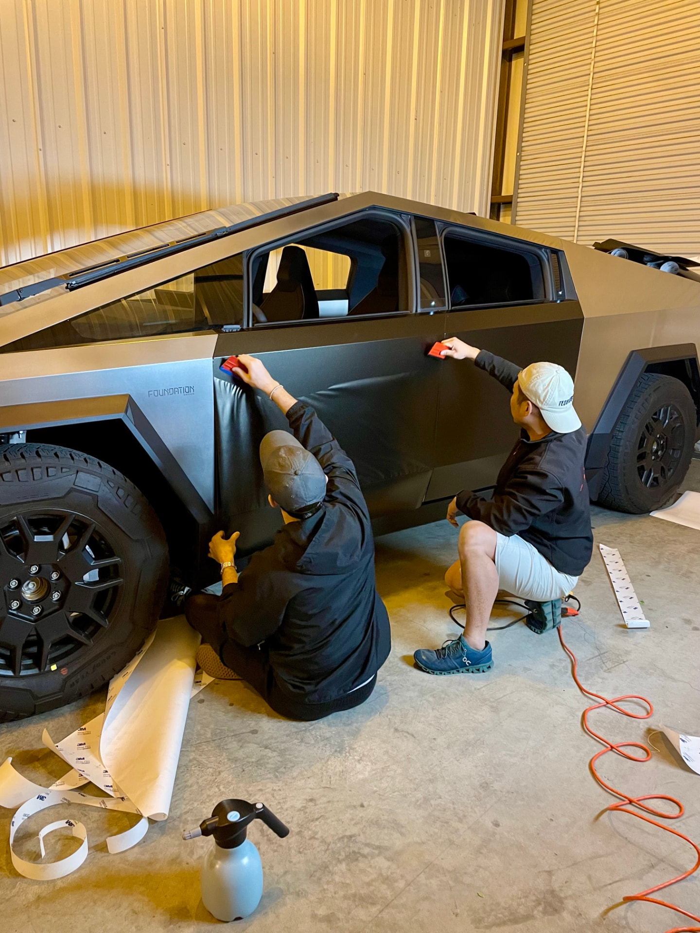 Two installers applying a black wrap panel to a vehicle in a workshop